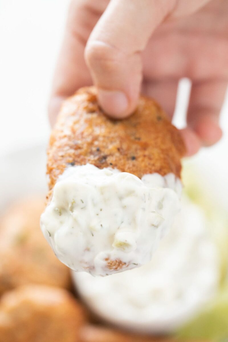 A hand with a beer-battered vegan fish stick with some vegan tartar sauce.