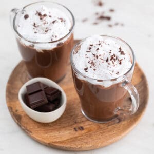 Two jars of vegan hot chocolate topped with vegan whipped cream and chopped dark chocolate.