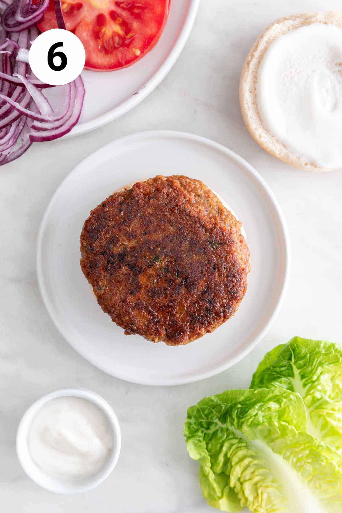 Vegan chickpea burger on a bun on a white plate with surrounding toppings.
