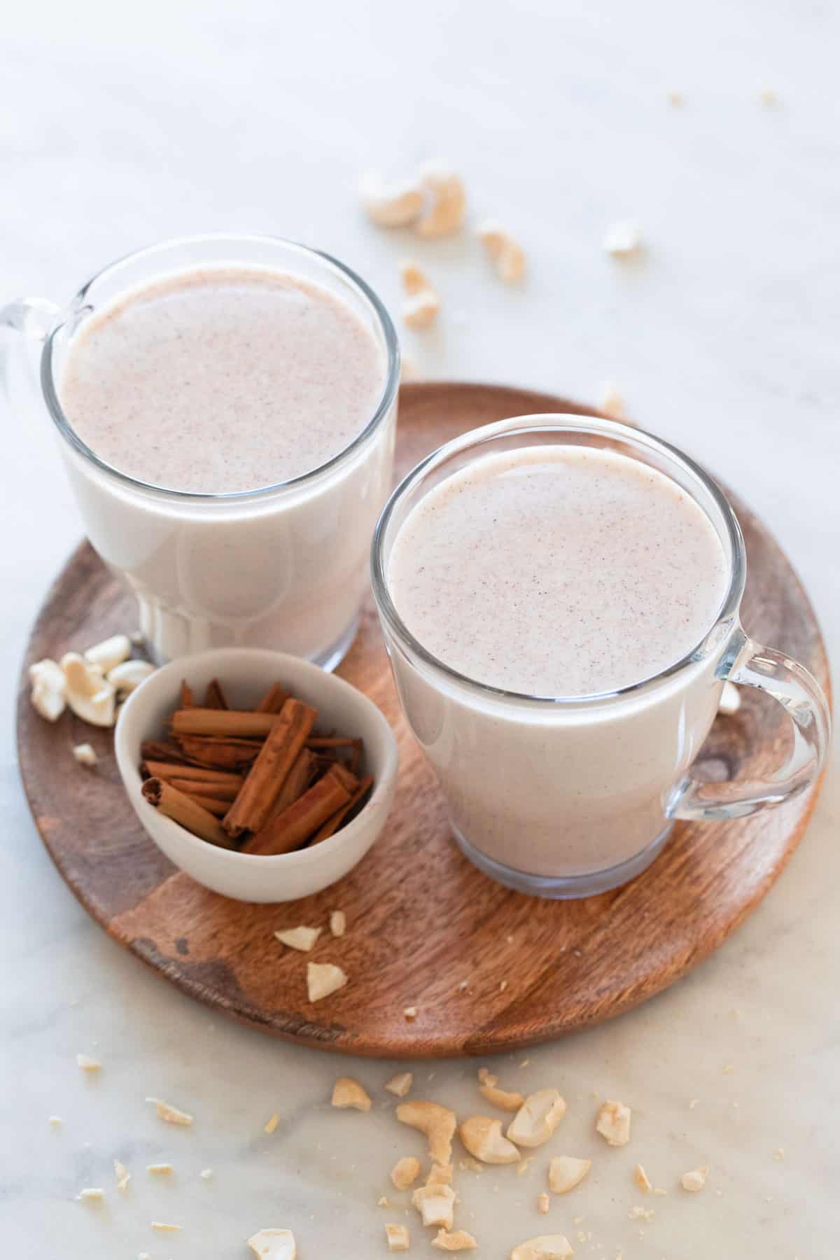 Two mugs of vegan eggnog onto a wooden board, a little bowl with cinnamon sticks and some cashews around.