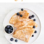 Overview of a plate with several folded vegan crepes topped with fresh blueberries and powdered sugar.