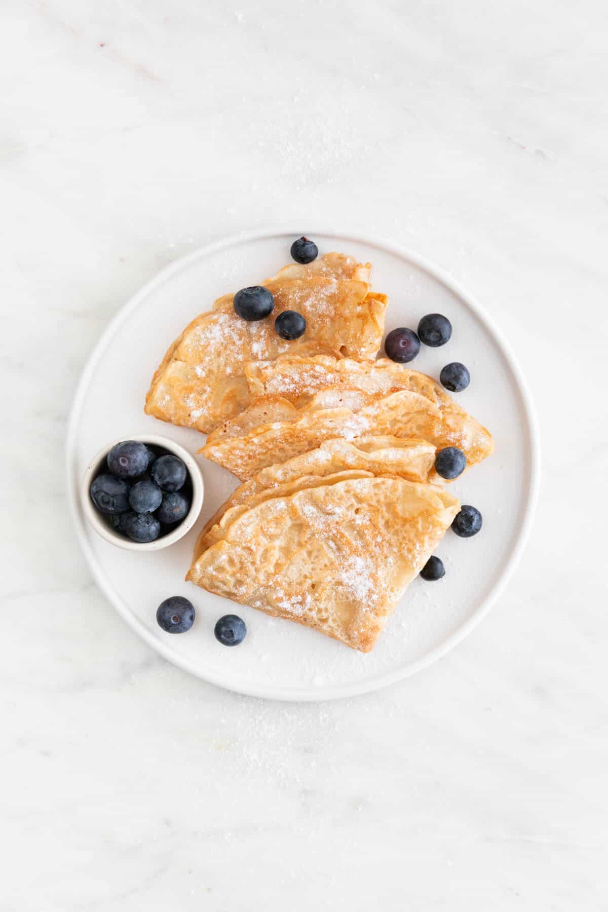 Overview of a plate with several folded vegan crepes topped with fresh blueberries and powdered sugar.