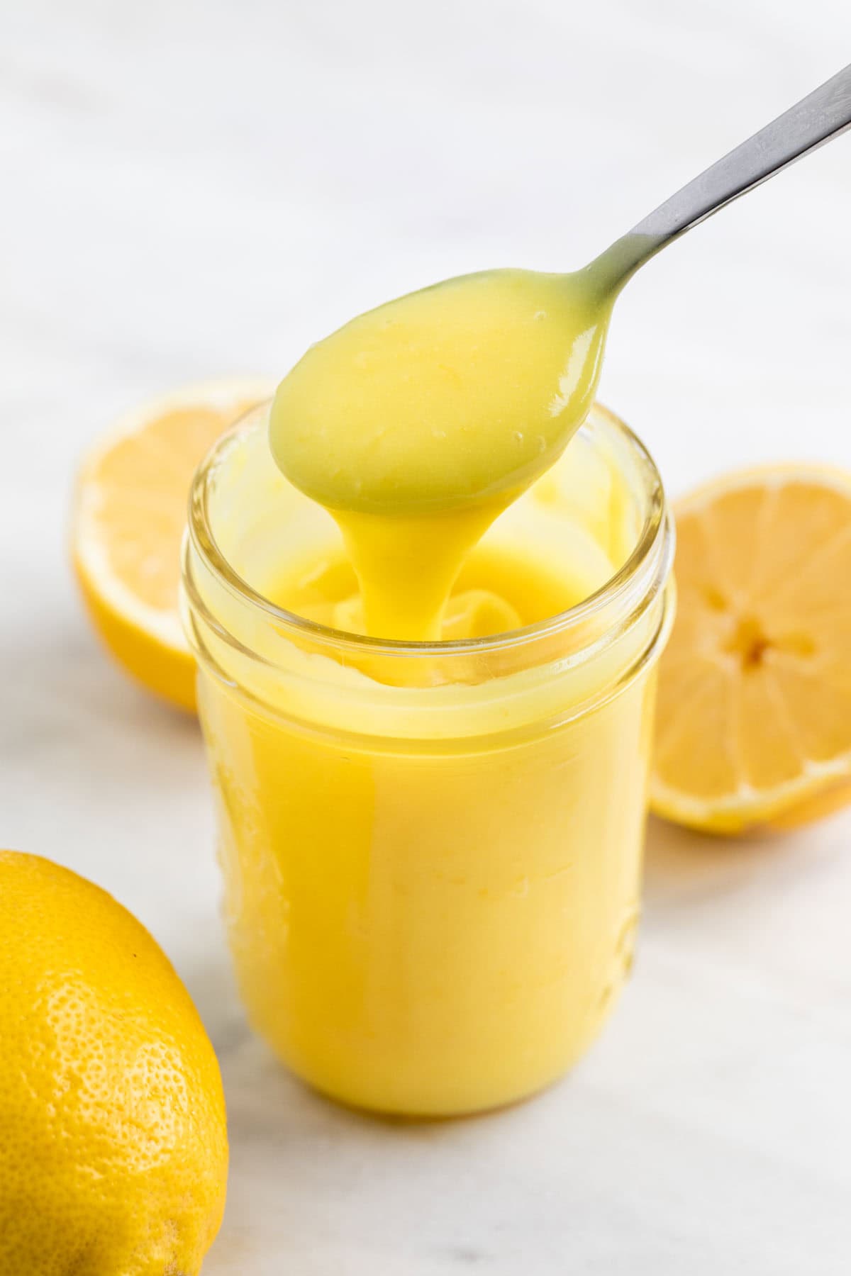 Jar of vegan lemon curd with lemons around and a spoon scooping curd from the jar.