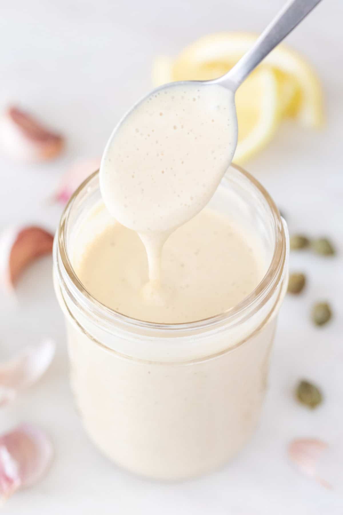 Mason jar with vegan Caesar dressing and ingredients in the background, being drizzled with a spoon.