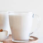 Two mugs of vegan eggnog onto a wooden board with some cashews and cinnamon sticks around.