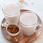 Two mugs of vegan eggnog onto a wooden board, a little bowl with cinnamon sticks and some cashews around.