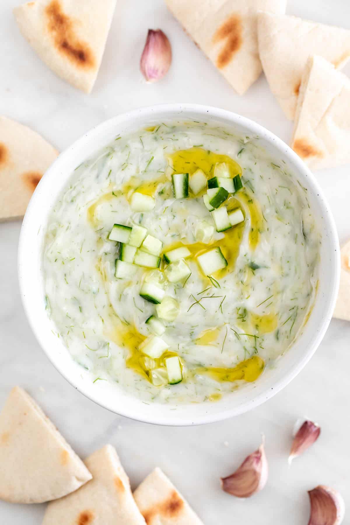 Bowl of vegan tzatziki sauce with pita bread and cloves of garlic in the brackground.
