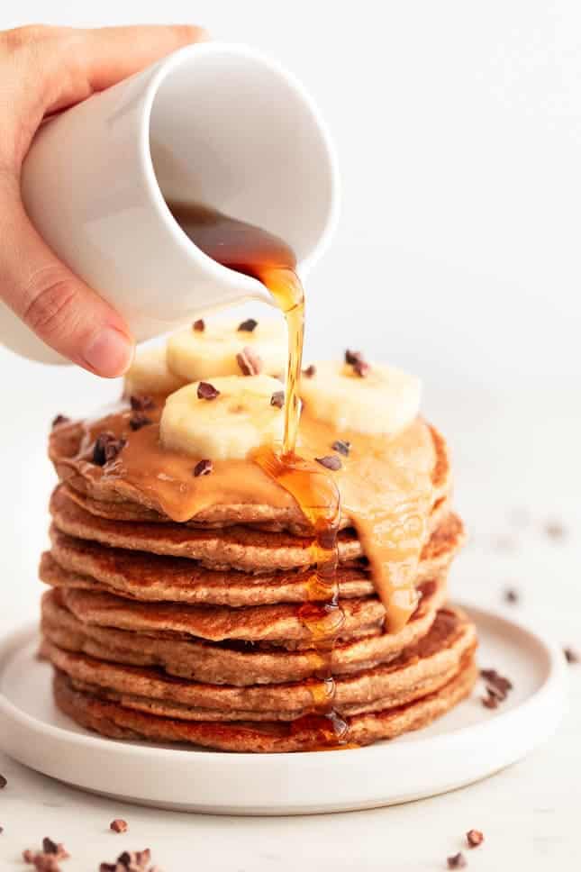 Stack of vegan banana pancakes topped with peanut butter, chocolate chips, banana, and syrup.