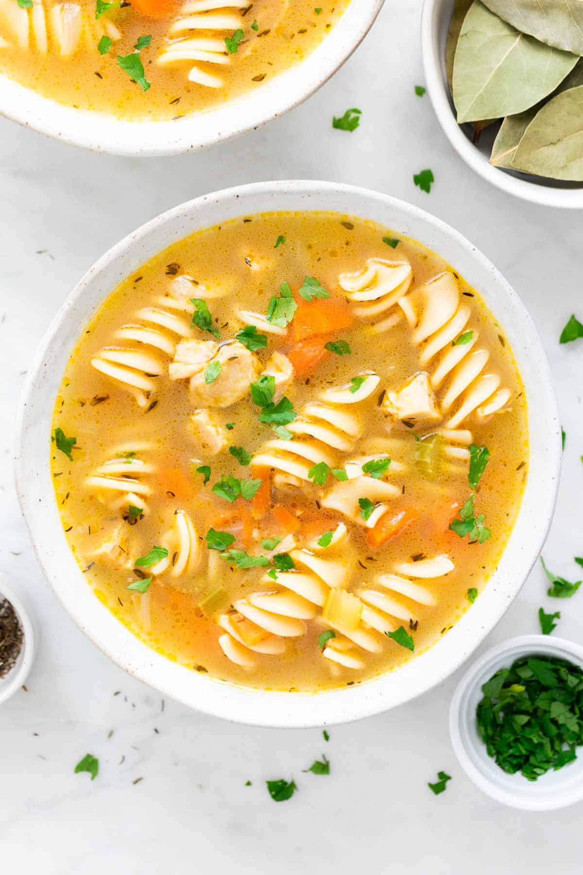 Two bowls of vegan chicken noodle soup garnished with parsley, surrounded by ingredients.