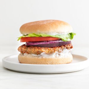 On a white plate, a vegan chickpea patty on a bun with lettuce, tomato, red onion, and mayo.