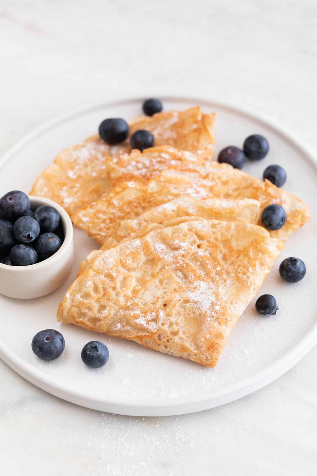 A plate with several folded vegan crepes with some blueberries and powdered sugar on top.