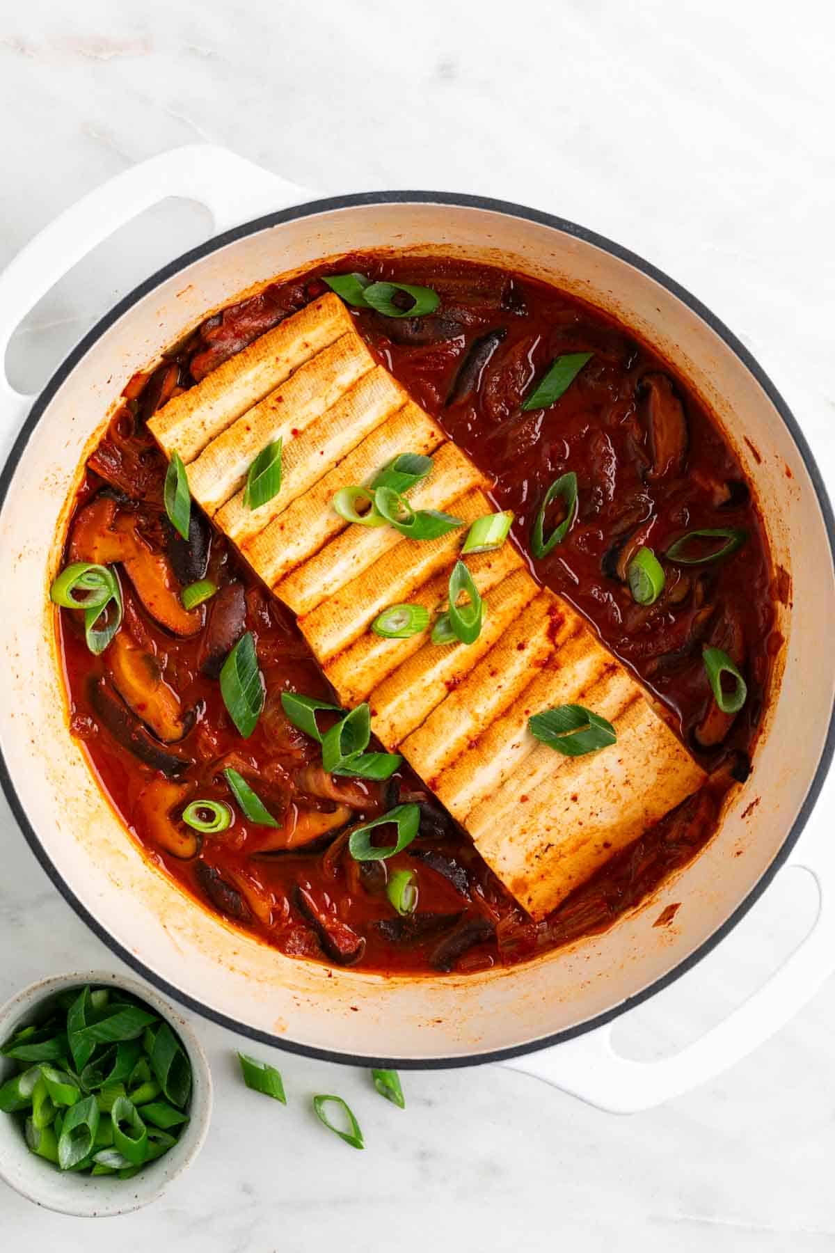 Vegan kimchi stew in a large pot with scallions on top, and a bowl of extra scallions on the side.