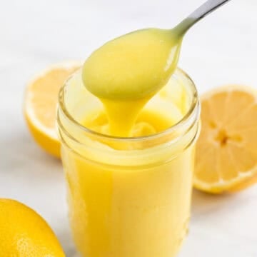 Jar of vegan lemon curd with lemons around and a spoon scooping curd from the jar.