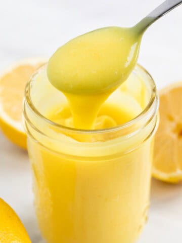 Jar of vegan lemon curd with lemons around and a spoon scooping curd from the jar.