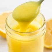 Jar of vegan lemon curd with lemons around and a spoon scooping curd from the jar.
