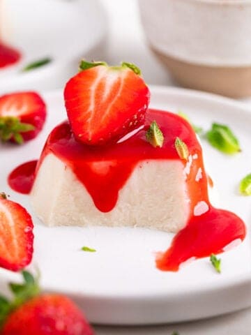 Vegan panna cotta on a plate, cut to reveal the inside, topped with strawberry coulis, a strawberry, and mint leaves.
