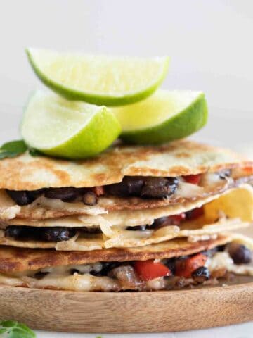 Side photo of a pile of vegan quesadillas onto a wooden board with some lime wedges
