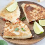 Photo of 3 pieces of vegan quesadilla onto a wooden board with some chopped cilantro and lime wedges with a heading