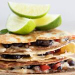 Side photo of a pile of vegan quesadillas onto a wooden board with some lime wedges and a heading