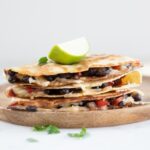 Square photo of a pile of vegan quesadillas onto a wooden board with a lime wedge