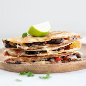 Square photo of a pile of vegan quesadillas onto a wooden board with a lime wedge