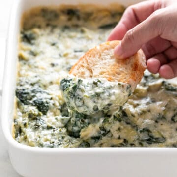 Hand dipping bread into vegan spinach artichoke dip in a baking dish.