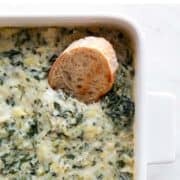 Baking dish with vegan spinach artichoke dip and a slice of bread inside.