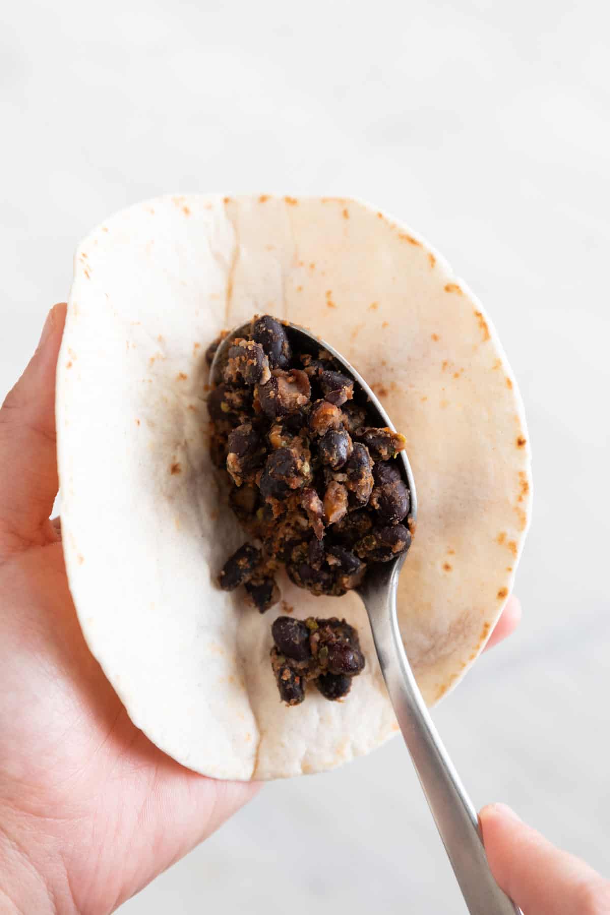 A hand with a tortilla and another hand with a spoon filling the tortilla with the black bean filling.