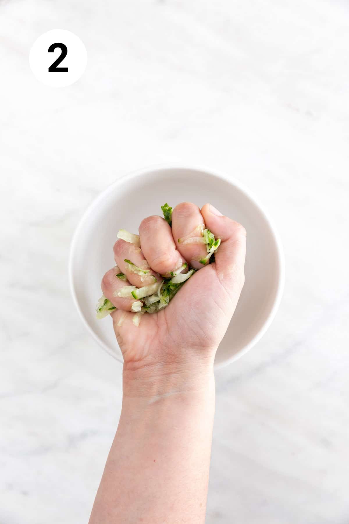 Hand squeezing out excess liquid from the grated cucumber over a bowl.